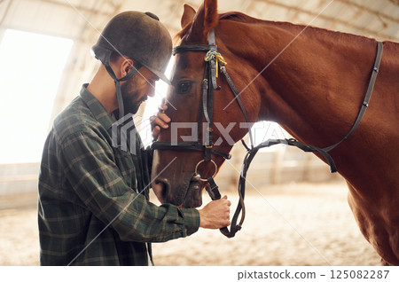 Standing and holding animal. Young man with a horse is in the hangar 125082287