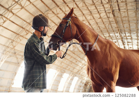 Standing and holding animal. Young man with a horse is in the hangar 125082288