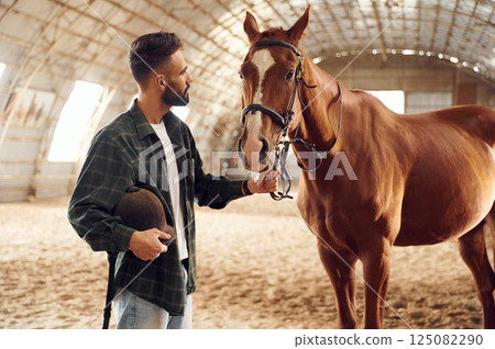 Standing and holding animal. Young man with a horse is in the hangar 125082290
