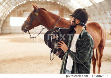 Holding the saddle in hands. Young man with a horse is in the hangar Holding the saddle in hands. Young man with a horse is in the hangar 125082306