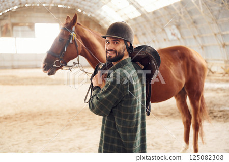 Holding the saddle in hands. Young man with a horse is in the hangar Holding the saddle in hands. Young man with a horse is in the hangar 125082308
