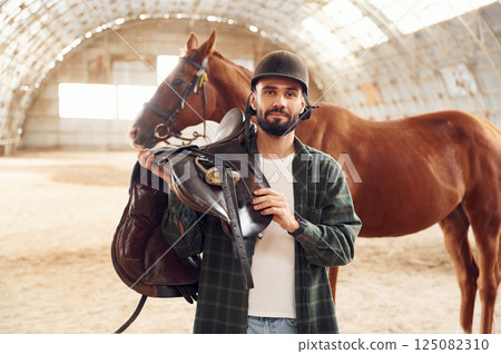 With saddle in hands. Young man with a horse is in the hangar With saddle in hands. Young man with a horse is in the hangar 125082310