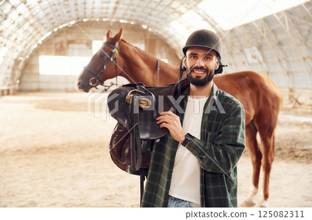 With saddle in hands. Young man with a horse is in the hangar With saddle in hands. Young man with a horse is in the hangar 125082311