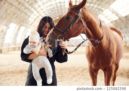 Standing and smiling. Mother with baby on the hands is indoors with horse in hangar 125082598