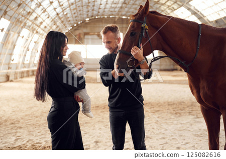 Lovely couple is standing with indoors in the hangar with little baby and showing him horse Lovely couple is standing with indoors in the hangar with little baby and showing him horse 125082616