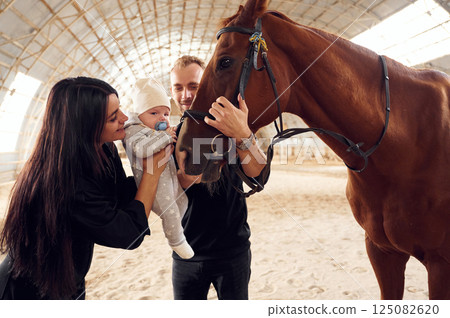 Lovely couple is standing with indoors in the hangar with little baby and showing him horse Lovely couple is standing with indoors in the hangar with little baby and showing him horse 125082620