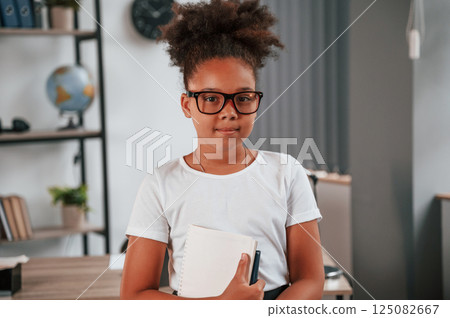 Holding paper notepad in hands. Cute african american child is standing in the domestic room Holding paper notepad in hands. Cute african american child is standing in the domestic room 125082667
