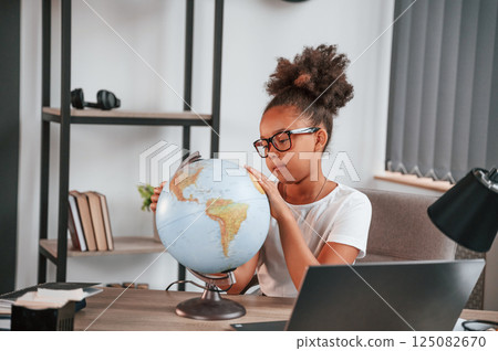 Examining the globe. Cute female african american student child is sitting by the laptop Examining the globe. Cute female african american student child is sitting by the laptop 125082670