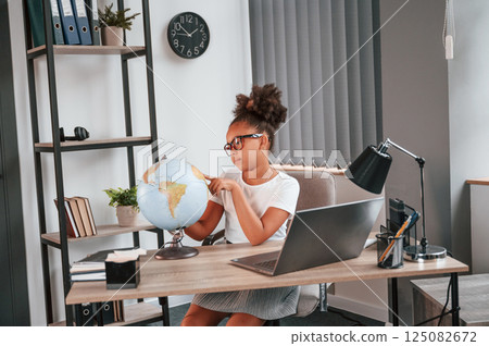 Examining the globe. Cute female african american student child is sitting by the laptop Examining the globe. Cute female african american student child is sitting by the laptop 125082672
