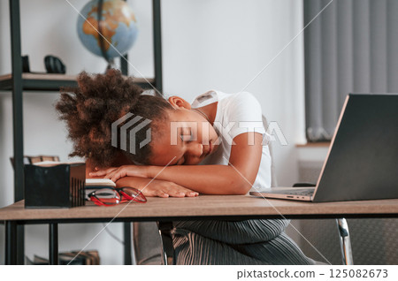 Sleeping by leaning on the table. Feeling tired. Cute female african american student child is sitting by the laptop 125082673
