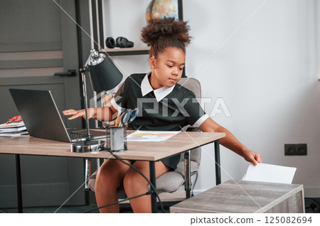 Holding paper. Sitting by the table with laptop. Cute female african american student child 125082694