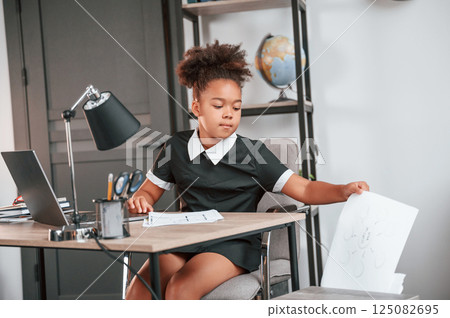 Holding paper. Cute female african american student child is sitting by the laptop Holding paper. Cute female african american student child is sitting by the laptop 125082695
