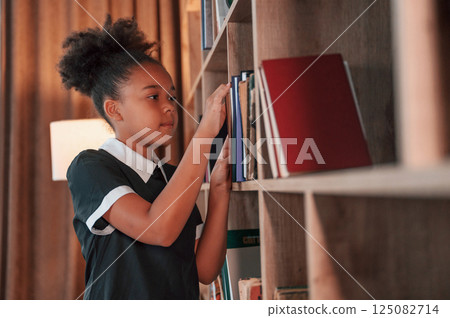 Searching for the books on the shelves. Cute african american girl in school uniform is at home library 125082714