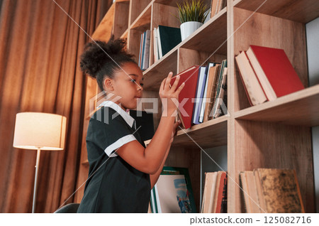 Searching for the books on the shelves. Cute african american girl in school uniform is at home library Searching for the books on the shelves. Cute african american girl in school uniform is at home library 125082716