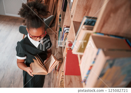Standing and reading the book. Cute african american girl in school uniform is at home library 125082721