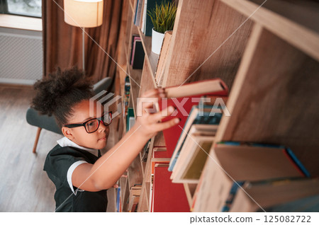 Searching for the books on the shelves. Cute african american girl in school uniform is at home library 125082722