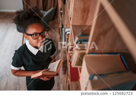 Searching for the books on the shelves. Cute african american girl in school uniform is at home library Searching for the books on the shelves. Cute african american girl in school uniform is at home library 125082723