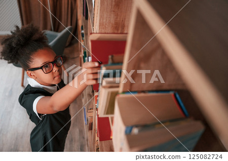 Taking the book from shelves. Cute african american girl in school uniform is at home library Taking the book from shelves. Cute african american girl in school uniform is at home library 125082724