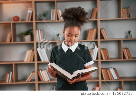Standing and reading the book. Cute african american girl in school uniform is at home library Standing and reading the book. Cute african american girl in school uniform is at home library 125082733