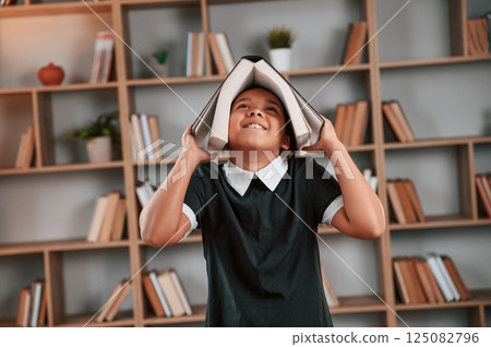 Having fun, book is on the head. Cute african american girl in school uniform is at home library Having fun, book is on the head. Cute african american girl in school uniform is at home library 125082796