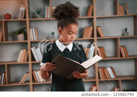 Holding book and reading. Cute african american girl in school uniform is at home library 125082799