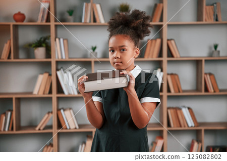 Holding big book. Cute african american girl in school uniform is at home library 125082802
