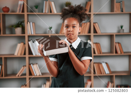 Holding big book. Cute african american girl in school uniform is at home library 125082803