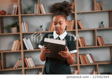 Holding big book. Cute african american girl in school uniform is at home library 125082804