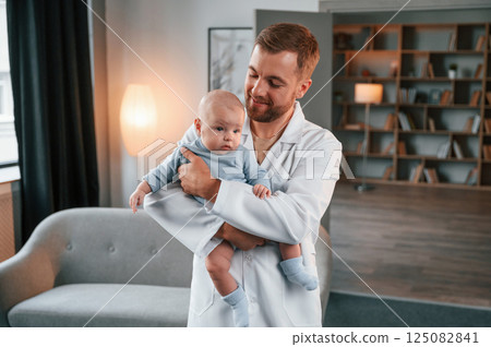 Looking at the kid. Doctor standing and holding newborn baby in hands indoors at apartment Looking at the kid. Doctor standing and holding newborn baby in hands indoors at apartment 125082841