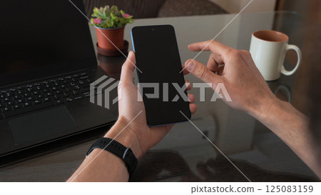 Man Holding Smartphone by Laptop in Modern Workspace Environment 125083159