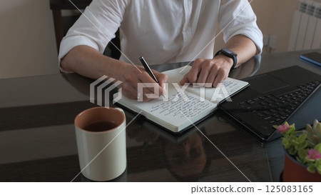 Man Writing Notes in Journal While Working with Laptop in Office Man Writing Notes in Journal While Working with Laptop in Office 125083165