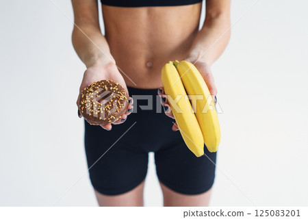 Sporty woman with a perfect body holding donut and bananas. Healthy low calorie meals, fresh fruits Sporty woman with a perfect body holding donut and bananas. Healthy low calorie meals, fresh fruits 125083201