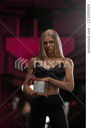 fitness model displays a muscular physique while holding a small container dressed in a fashionable athletic outfit. 125085926