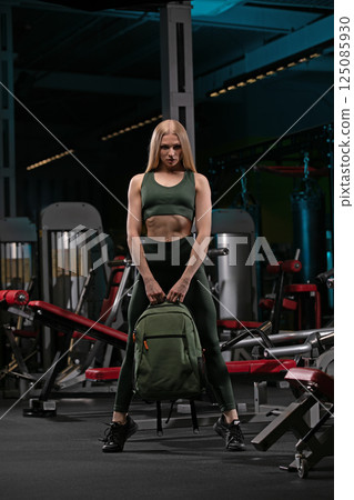 sporty woman in a green crop top and leggings is striking a confident pose in a well equipped gym. She holds a backpack in front of her showcasing her strength and fitness dedication. sporty woman in a green crop top and leggings is striking a confident pose in a well equipped gym. She holds a backpack in front of her showcasing her strength and fitness dedication. 125085930