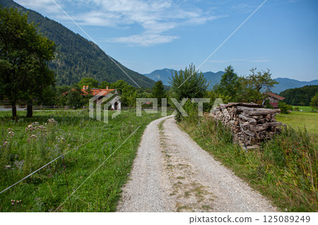 Serene gravel path winding through lush fields and towering mountains in a picturesque German countryside 125089249