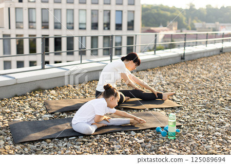 Middle-aged woman and young girl practicing stretching exercises on rooftop yoga mats outdoors 125089694