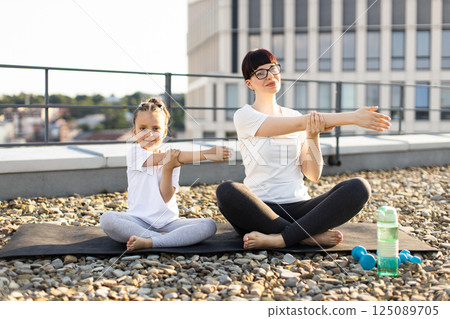 Female adult and young girl doing fitness yoga stretches outdoors. Caucasian family enjoying healthy sports activity under clear sky on city rooftop. Female adult and young girl doing fitness yoga stretches outdoors. Caucasian family enjoying healthy sports activity under clear sky on city rooftop. 125089705