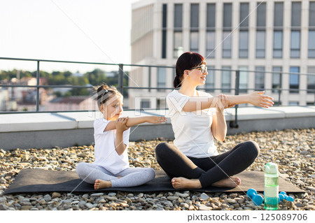 Adult woman and young girl seated outdoors wearing activewear doing arm stretches on rooftop. Image conveys healthy lifestyle, family connection, fitness, calmness, and outdoor activity in warm light. 125089706