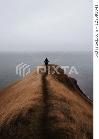 Person standing on grassy hill slope overlooking ocean under cloudy sky 125093041