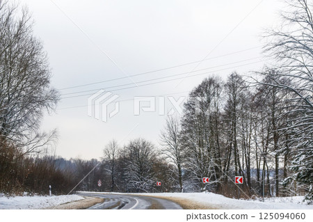 Snow-covered winter road with visible traffic signs warning of slippery conditions. The cold, icy atmosphere highlights the challenges of driving in winter weather, with snow blanketing 125094060
