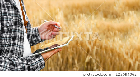 Modern farmer standing in a golden wheat field during summer, using a tablet to monitor crops. Concept of agribusiness, smart farming, and agricultural technology integration. Copy space 125094062