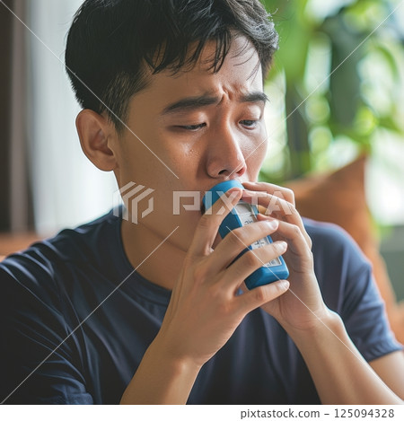 A young man uses an inhaler, experiencing respiratory distress indoors 125094328