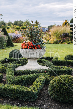 Shaped interlacing green hedges surrounding the base of a round stone pedestal in a landscaped garden. Garden design, sustainable horticulture, biodiversity integration, geometric landscaping Shaped interlacing green hedges surrounding the base of a round stone pedestal in a landscaped garden. Garden design, sustainable horticulture, biodiversity integration, geometric landscaping 125095110