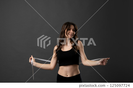 Beautiful brunette young woman in black outfit with measuring tape. Measuring tape on the waist. Beautiful fitness woman is in the studio. 125097724
