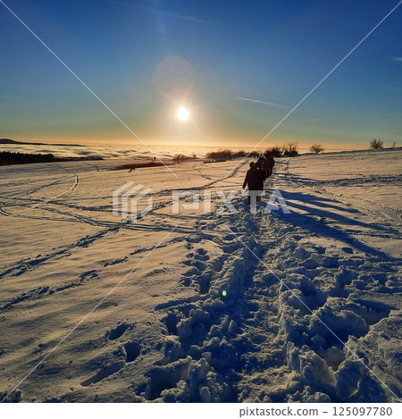 The concept of relaxing in the mountains in winter in the snow on skis, snowboards or sleds, walking under the setting sun at sunset on the Wasserkuppe mountain in Hesse Germany 125097780