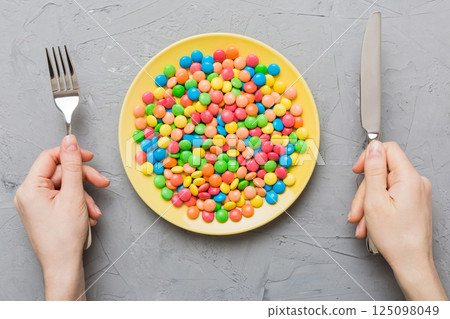 the girl holds cutlery in her hands and eats sweets in a plate. Health and obesity concept, top view on colored background the girl holds cutlery in her hands and eats sweets in a plate. Health and obesity concept, top view on colored background 125098049