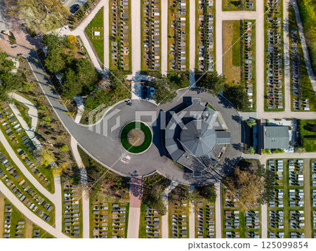 Aerial view of a cemetery with symmetrical rows of graves, a central round building, and surrounding greenery. Pathways and trees create an organized, peaceful layout from above. Aerial view of a cemetery with symmetrical rows of graves, a central round building, and surrounding greenery. Pathways and trees create an organized, peaceful layout from above. 125099854