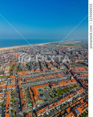 A stunning aerial view of a coastal town with red-tiled rooftops, neatly aligned houses, and a vast sandy beach stretching into the distance under a clear blue sky. 125099858