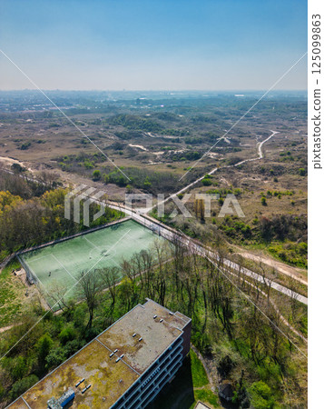 Aerial view of a football field near a building, surrounded by trees and trails, with vast open hills and distant cityscape under a clear blue sky. 125099863