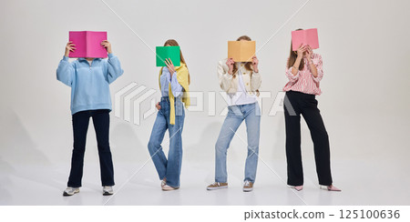 Group of students pose with colorful books covering faces, standing casually in modern classroom on white background. Group of students pose with colorful books covering faces, standing casually in modern classroom on white background. 125100636